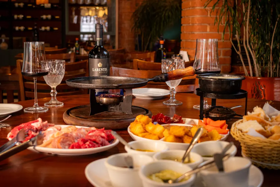 Mesa de restaurante com fondue de queijo, chapa de ferro, carnes, pães, molhos e taças de vinho tinto.