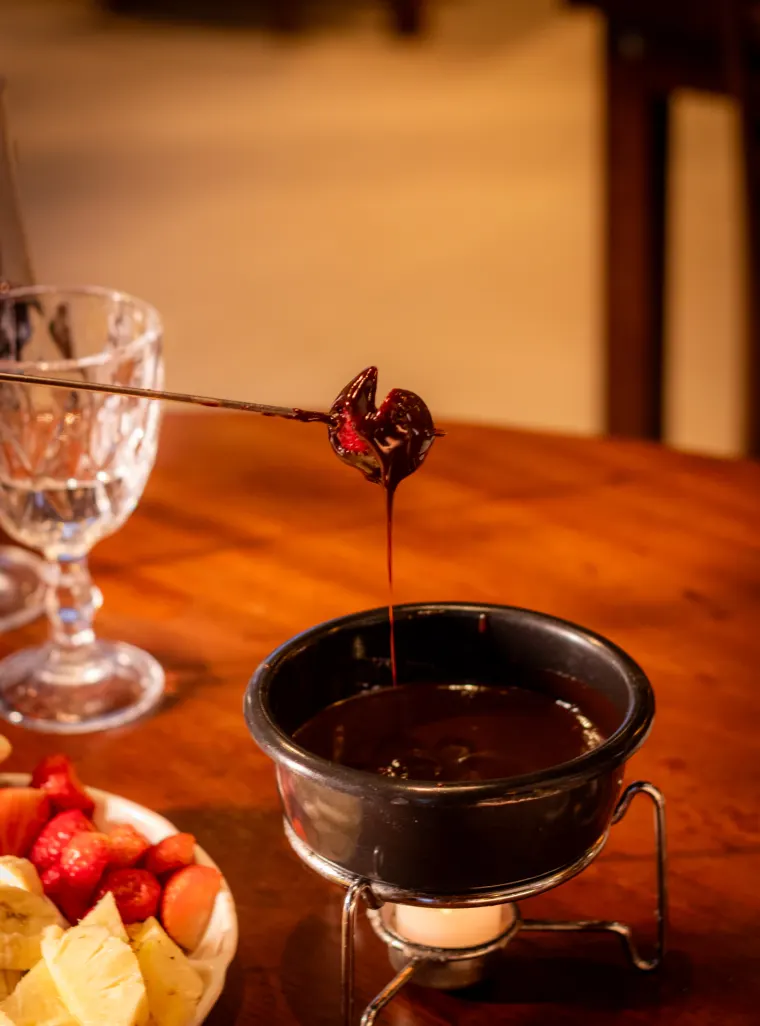 Morango coberto de chocolate escorrendo sobre a panela de fondue doce, com frutas picadas e uma taça ao lado.