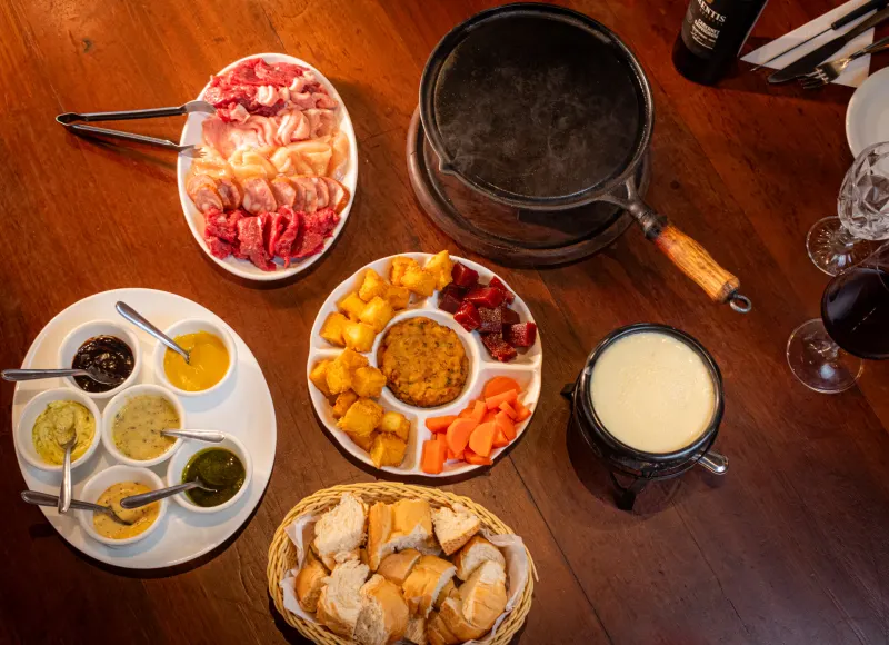 Mesa de fondue vista de cima: panela de queijo, chapa de ferro, carnes, molhos, polenta frita e cesta de pães.