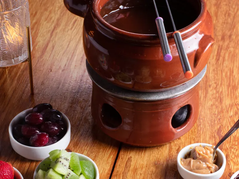 Fondue de chocolate em panela de cerâmica, com potes de uva, kiwi, morango, goiabada e bolo.