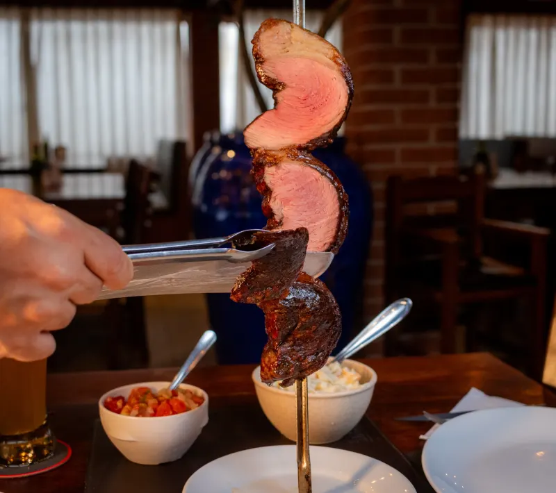 Picanha no espeto sendo fatiada à mesa, com vinagrete e maionese servidos ao lado.