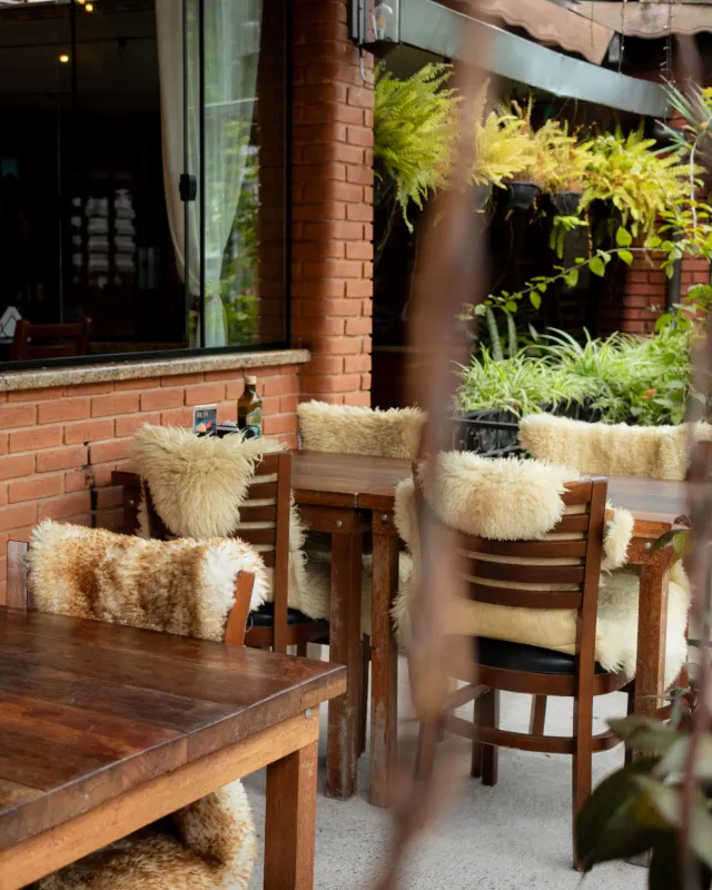 Varanda de restaurante com mesas de madeira, cadeiras cobertas com pelegos e parede de tijolos, cercada de plantas.